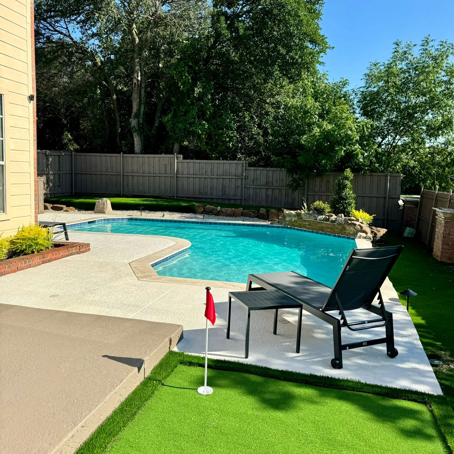 JackontOutdoor Backyard pool with lawn chairs, a small putting green, and trees and fence in the background on a sunny day.