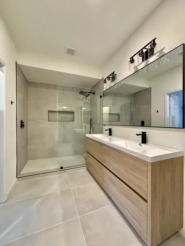 bathroom-2 Modern bathroom with glass shower, double sink with wooden vanity, large mirror, and neutral tile flooring.