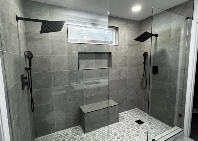 Modern double shower with gray tiles, glass doors, black fixtures, a bench, and hexagonal floor tiles.