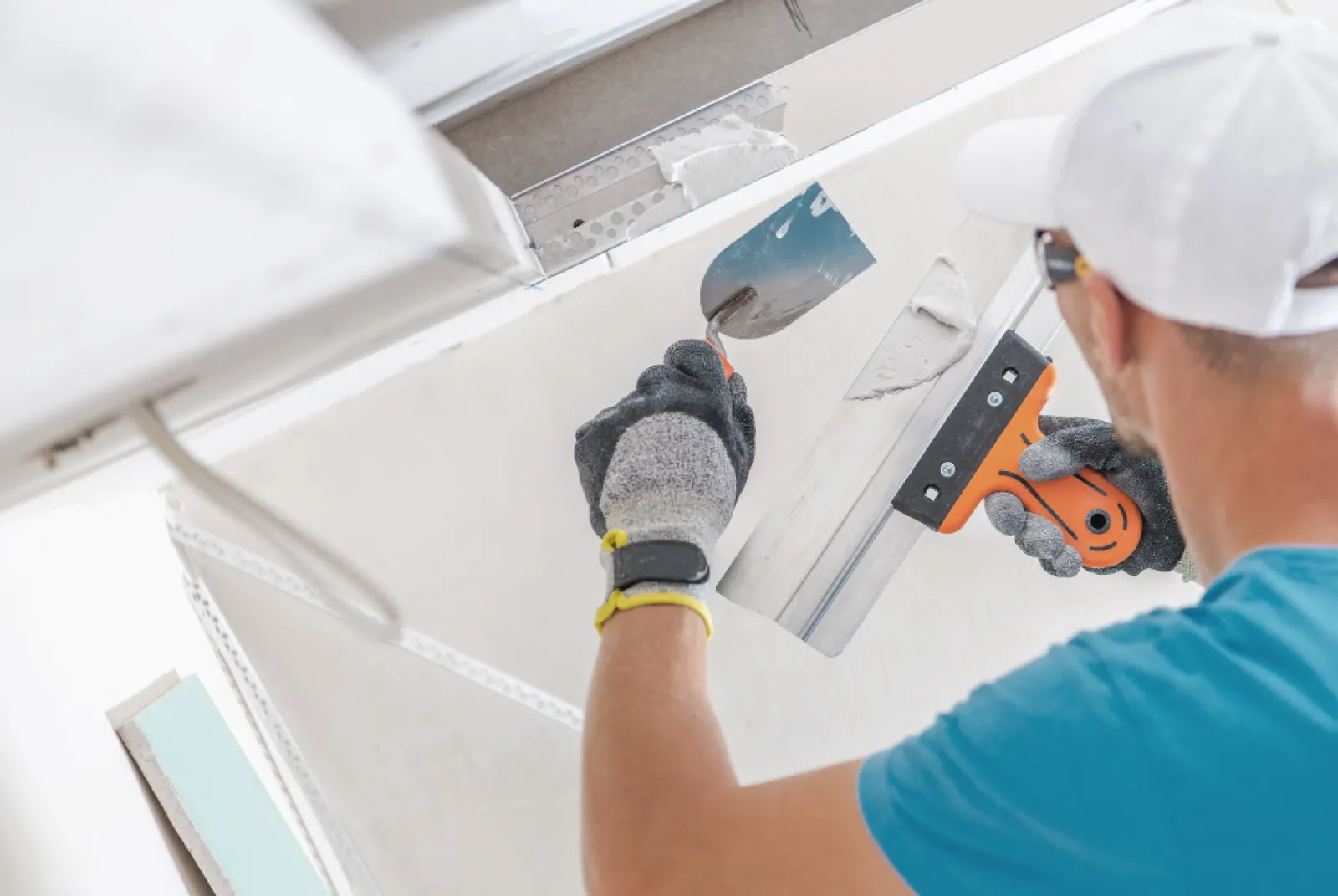 Worker in gloves and cap cuts a hole in a white drywall ceiling using a utility knife and a metal tool.
