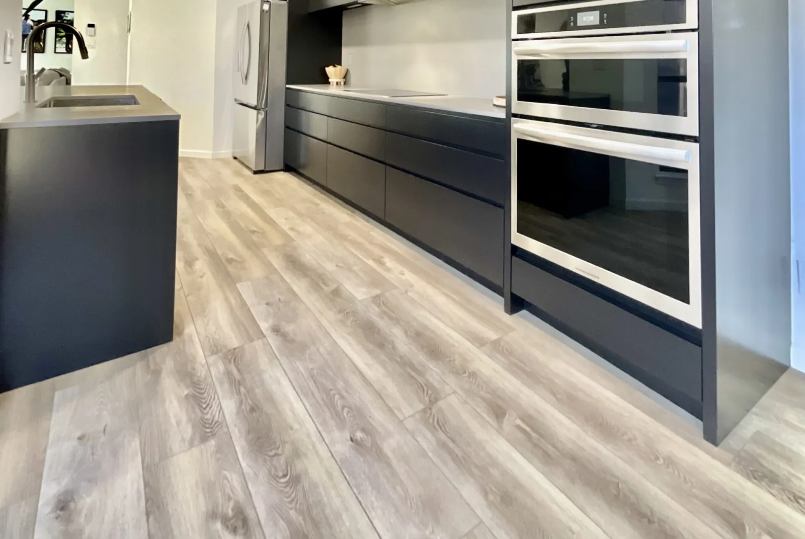 flooring-1 Modern kitchen with light wood flooring, black cabinets, stainless steel appliances, and a gray backsplash.