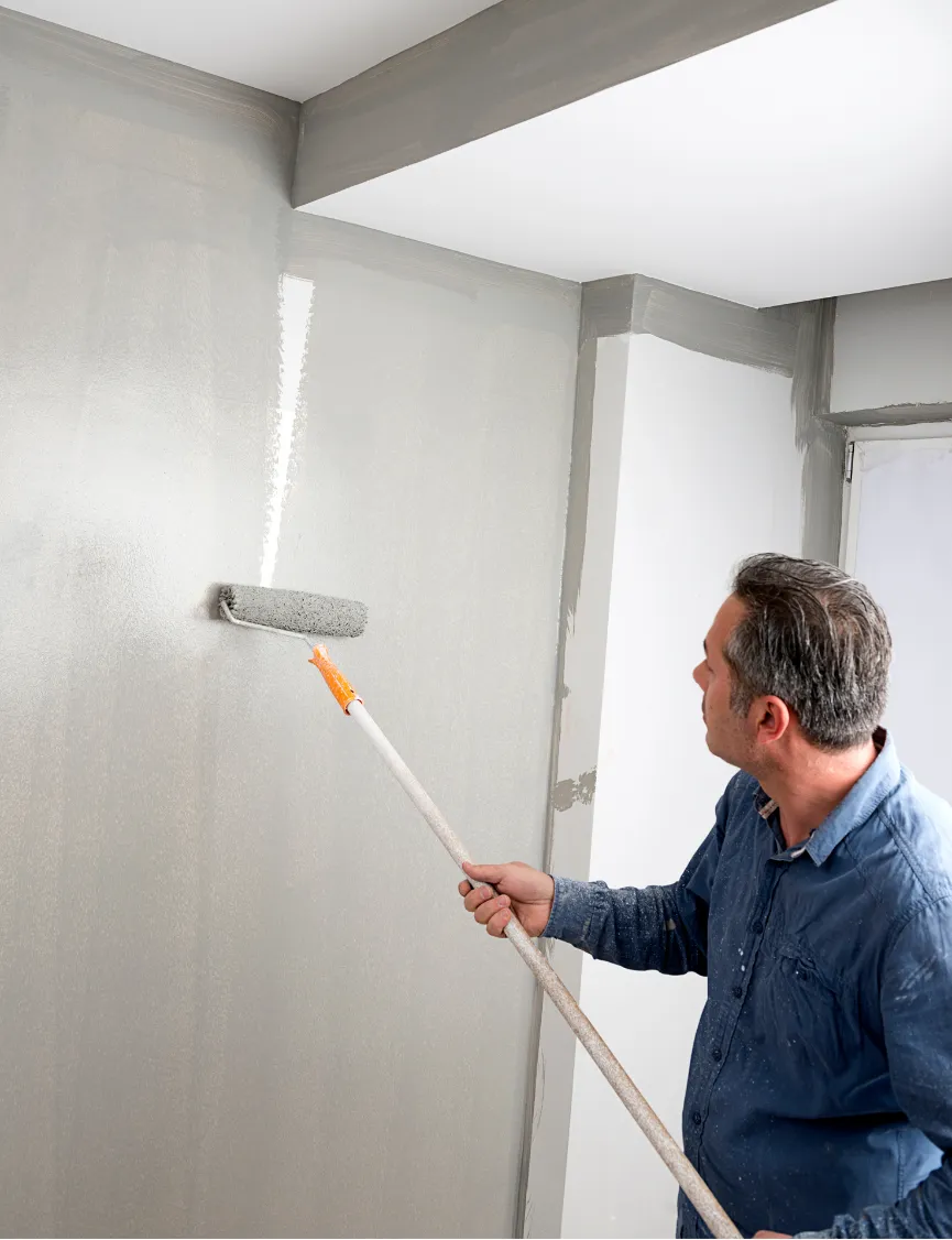 painting-1 A man painting a wall light gray with a roller on an extension pole inside a room.