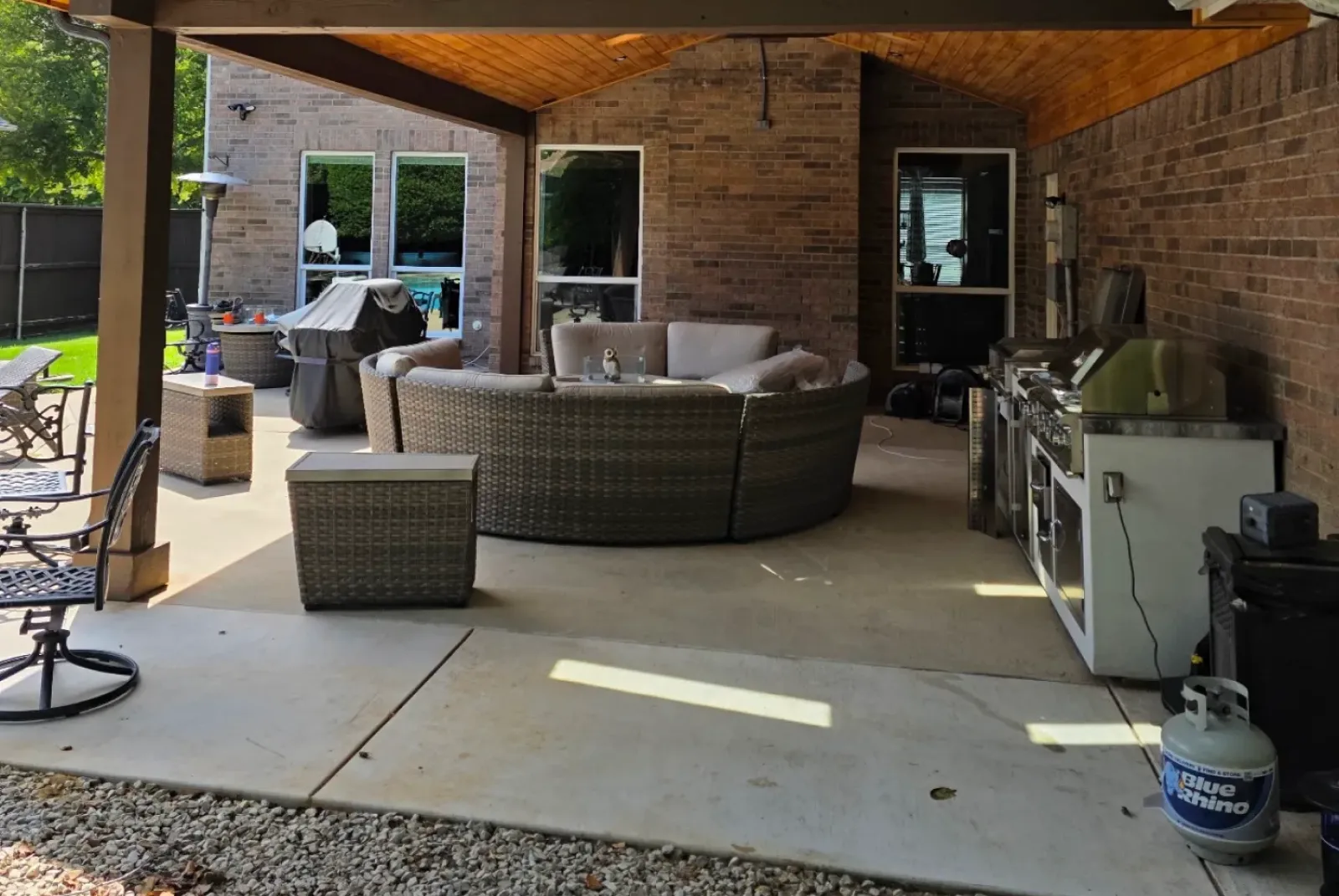 Covered patio with round wicker sofa set, grill, and outdoor kitchen against a brick wall.