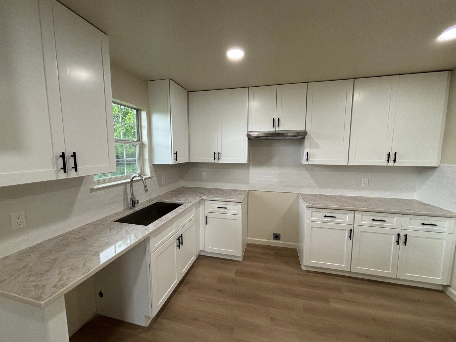 Modern kitchen with white cabinets, marble countertops, and wood flooring under bright overhead lighting.