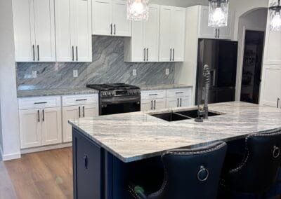 Modern kitchen with white cabinets, marble backsplash, black appliances, and a navy island with bar stools.
